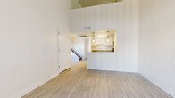 This well-designed unit offers a clean, light-filled interior defined by its striking vaulted ceilings that lend a sense of openness and airiness to the main living area. Light-toned wood-style flooring runs throughout, complementing the bright white walls and creating a cohesive, modern aesthetic.

The open-concept living area connects seamlessly to a kitchen pass-through, which features pendant lighting and a countertop bar that serves as both a functional workspace and a casual dining surface. The galley-style kitchen is efficiently laid out with ample white cabinetry, a stainless steel range, dishwasher, and refrigerator, as well as a double sink — providing everything needed for day-to-day meal preparation.

A secondary room showcases an exposed concrete block accent wall, adding a subtle industrial character, and is bathed in natural light from a large sliding glass door fitted with vertical blinds that opens to an outdoor area.

A carpeted staircase with a white metal railing leads to additional levels of the building, visible from the main entryway. The front entry area is generous and transitions smoothly into the living space. Overall, the unit maintains a clean, neutral palette throughout, creating a versatile and inviting atmosphere ready for personalization.