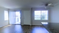 This single-story space showcases a clean, functional design centered around an open living area. The flooring throughout is a warm wood-style hardwood that adds character and visual continuity to the space. Neutral gray walls provide a versatile backdrop that complements a variety of décor styles.

Natural light is a defining feature of this space, streaming in through a large picture window and a sliding glass door fitted with vertical blinds, both of which face an outdoor area visible through the glass. The sliding door offers convenient direct access to the exterior. A ceiling fan is mounted overhead, providing both air circulation and a subtle design element.

The front entry is anchored by a solid white door with a secure multi-point lock, offering a clean and welcoming focal point. A granite or stone-surfaced counter is partially visible along one edge of the space, suggesting a kitchen or bar area in close proximity to the main living room.

Overall, the space feels bright and airy, with a straightforward, well-maintained interior that balances practicality with a comfortable, livable aesthetic suited for everyday use.