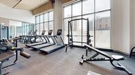 Located in Hamilton, Ontario, this contemporary fitness facility offers a thoughtfully designed workout space that combines striking architectural elements with a comprehensive range of exercise equipment. The space is defined by soaring double-height ceilings adorned with a distinctive slatted wood panel design accented by bold burgundy beams, creating a visually dynamic overhead feature that sets the tone for the entire room.

Floor-to-ceiling windows line one side of the facility, flooding the space with abundant natural light and offering views of the surrounding Hamilton cityscape. The open-concept layout is covered in durable rubber flooring throughout, providing both safety and functionality for all types of exercise.

The cardio zone features a row of modern treadmills and stationary bikes equipped with digital screens, while the strength training area is outfitted with a Smith machine, cable systems, a multi-station weight machine, adjustable benches, and a free weights rack. Spin bikes are positioned near a mirrored wall, which visually expands the space and enhances the sense of openness.

Large glass partitions contribute to the airy, connected feel of the facility, while recessed lighting supplements the generous natural light. The overall ambiance is clean, modern, and energizing, making this fitness centre a well-equipped and inviting space for a full-body workout.
