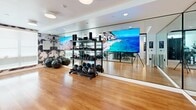 Located at 1972 First St in Livermore, California, this contemporary fitness center offers a thoughtfully designed workout environment across a single floor. The space is divided into two distinct functional areas, seamlessly connected by sleek glass-panel partitions that lend an open, airy feel to the overall layout.

The main gym area features polished hardwood flooring and is lined with floor-to-ceiling mirrors that visually expand the space. Large-format display screens are mounted along the mirrored wall, providing an immersive visual backdrop during workouts. The room is well-equipped with a comprehensive selection of free weights, including kettlebells, medicine balls, and stability balls, all neatly organized on dedicated racks. Recessed and flush-mount ceiling lighting ensures consistent, bright illumination throughout.

Adjacent to the main floor, a separate section houses a variety of professional-grade strength and resistance machines, set atop light-toned carpet flooring. This area is accessible through glass doors, maintaining visual connectivity while offering a distinct training environment.

A smaller studio room, also accessible via a black-framed glass sliding door, features warm hardwood floors and striking abstract-patterned accent wallpaper. Roller shades on the windows allow for adjustable natural light, making it well-suited for yoga, stretching, or group fitness sessions. Together, these spaces form a cohesive, modern fitness facility with high-end finishes and versatile training options.