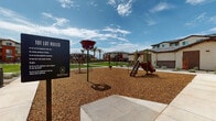 Located at 7430 N 100th Ave in Glendale, Arizona, this outdoor playground is part of the Desert River residential community. The play area is thoughtfully designed with safety and accessibility in mind, featuring a generous wood chip surface that cushions the ground throughout the entire space. Multiple play structures are spread across the area, including a multi-level climbing structure with a ladder and dual slides, interactive spinning and sensory play elements, and additional freestanding equipment that caters to a range of ages and abilities. Concrete walkways with tactile detailing border the perimeter, ensuring accessible entry and circulation around the playground. A decorative shade structure with a laser-cut canopy provides a covered retreat at the edge of the play zone, complemented by desert-adapted landscaping including flowering shrubs and native plantings. Tall palm trees and patches of green turf are visible in the background, adding a lush contrast to the Arizona desert setting. The surrounding Desert River community buildings — featuring a contemporary Southwest architectural style with stucco exteriors and earth-tone accents — frame the space on multiple sides. Community rules signage is prominently posted at the entrance, and the open layout allows for clear sightlines across the entire play area, making it easy to supervise children throughout.