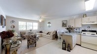 This single-floor home offers an open-concept design that creates a welcoming and cohesive living environment. The main living area is generously proportioned, featuring large windows that flood the space with abundant natural light and offer views of the greenery outside. A ceiling fan overhead adds both a functional and decorative touch to the bright, white ceiling. The living room is anchored by a comfortable sectional sofa and complemented by stylishly patterned accent chairs, creating an inviting atmosphere for relaxation and entertaining. Light-toned tile flooring runs throughout, enhancing the sense of openness and easy flow between areas. Transitioning seamlessly from the living room, the kitchen features a peninsula countertop with a light stone surface, providing additional seating with bar-height chairs — an ideal spot for casual dining or conversation. White cabinetry lends a clean, classic look to the kitchen space. Decorative accents including framed artwork, a round mirror, lanterns, and fresh floral arrangements add warmth and personality throughout. The overall ambiance is light, neutral, and elegantly understated, making this a comfortable and visually appealing single-level home.