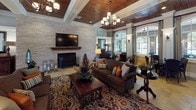 This sophisticated single-floor space showcases a refined blend of traditional Southern and transitional design elements, creating a warm and inviting atmosphere from the moment you enter. The grand foyer sets the tone with large-format travertine tile flooring, a stately arched entryway, decorative wainscoting, and a rich wood-paneled ceiling that carries throughout much of the space. Wall sconces and a dark wood console table with a framed mirror add an air of classic elegance to the entry.

The main living area is anchored by a striking whitewashed stone fireplace wall, complete with a wood mantle and mounted flat-screen television above. Coffered wood ceilings with recessed lighting and a chandelier overhead lend a sense of grandeur, while plush seating arranged around a carved wood coffee table invites relaxed gatherings. Patterned area rugs add warmth and texture to the travertine floors.

A casual dining area offers multiple seating vignettes with dark leather chairs surrounding round tables, all bathed in natural light pouring through a stunning series of floor-to-ceiling arched windows. These oversized windows frame views of an outdoor covered terrace, seamlessly connecting the interior to the exterior. Tall decorative columns, draped curtains, and lush potted plants further enhance the refined yet comfortable character of this beautifully appointed space.
