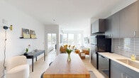 This thoughtfully designed single-floor unit offers a clean, modern aesthetic throughout its one bedroom and one bathroom layout. Light oak hardwood flooring runs continuously across all rooms, anchoring the bright, white-walled interior and creating a cohesive sense of flow.

The open-concept kitchen is outfitted with sleek gray cabinetry, white countertops, a tile backsplash, and black stainless steel appliances including a refrigerator, electric range, built-in microwave, and dishwasher. A butcher block kitchen island with bar seating provides additional prep space and a casual dining option.

The living room benefits from large, tall windows that frame outdoor views and flood the space with natural light. Recessed ceiling lighting complements the brightness, and the room comfortably accommodates a full seating arrangement.

The bedroom features a large window with outdoor views, a wall-mounted mini-split HVAC unit for climate control, and ample room for furnishings. The bathroom maintains the minimalist design language of the unit, with gray cabinetry, a white countertop vanity, and black hardware accents throughout.

A dedicated laundry area houses a stacked full-size washer and dryer, adding a practical and convenient amenity. Overall, the unit presents a cohesive modern design with efficient use of space and quality finishes.