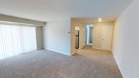 This single-floor unit offers a practical and comfortable layout designed for everyday living. The open living area is generously sized, finished with neutral-toned carpet and light gray walls that create a clean, understated aesthetic. A large sliding door with vertical blinds allows natural light to fill the space, enhancing its airy feel. A hallway connects the living area to the rest of the unit, with a stainless steel refrigerator visible at the transition point between the living and kitchen spaces.

The kitchen is efficiently arranged, featuring white cabinetry, white countertops, and a suite of stainless steel appliances including an electric range, over-the-range microwave, and dishwasher. A window above the sink brings in additional natural light, complemented by wood-look flooring that grounds the space.

The bedrooms are carpeted and neutrally painted, with one room notable for its dual closets — each illuminated and equipped with hanging rods — offering generous storage. A secondary bedroom provides a window with natural light and ample wall space.

The bathroom includes a full soaking tub and shower combination with cream-toned tile surround and brushed nickel grab bars and fixtures, adding both style and functionality to the space.