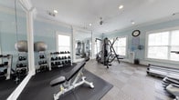 Located at 2222 Crow Ln in Myrtle Beach, South Carolina, this single-floor fitness center is a well-appointed exercise facility designed to support a full range of workout routines. The space is finished with soft blue walls, crisp white crown molding, and a tray ceiling accented by recessed lighting and a ceiling fan, creating a bright and airy atmosphere. Natural light pours in through multiple windows fitted with blinds, further enhancing the open feel of the room. The flooring features a combination of patterned carpet tiles and rubberized mat sections, providing both comfort and functionality for various exercise activities. The cardio zone is equipped with treadmills, elliptical machines, and a rowing machine, each positioned for easy use, with wall-mounted televisions nearby for entertainment during workouts. On the strength training side, a multi-station cable machine and an adjustable weight bench are available alongside a well-stocked rack of kettlebells and dumbbells spanning a wide range of weights. Large wall mirrors on one side of the room expand the visual space and allow users to monitor their form. A water cooler and informational postings round out the practical amenities, making this fitness center a comprehensive and inviting space for residents.