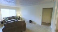 This single-floor space presents a functional and understated living area as its central feature. The room is finished with wall-to-wall carpeting in a light beige tone, contributing to a warm and quiet atmosphere underfoot. Neutral, cream-colored walls keep the space feeling open and adaptable to various furnishing styles. The ceiling features a textured finish, adding a subtle visual element overhead. Natural light enters through a window on one side of the room, brightening the space during daytime hours. The living area is generously sized, comfortably accommodating a full seating arrangement, as evidenced by the placement of a sofa and loveseat. A doorway on the far wall leads to additional areas of the home, suggesting a straightforward and accessible floor plan. The overall ambiance of the space is calm and neutral, offering a blank canvas that lends itself well to personalization. The simple, clean design prioritizes practicality, making it a versatile living environment suited to a range of lifestyles.