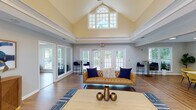 Located at 100 Eclipse Dr in Cary, NC, this single-story community clubhouse offers a thoughtfully designed gathering space with a variety of functional areas. The centerpiece of the building is a grand great room distinguished by soaring vaulted ceilings with an arched transom window that floods the space with natural light. French doors line the exterior walls, opening views to the surrounding greenery and providing seamless indoor-outdoor connectivity. Rich hardwood flooring runs throughout the main living areas, anchored by stylish area rugs and contemporary furnishings including tufted leather and upholstered sofas, accent chairs, and curated décor. Crown molding and a two-tone wall treatment lend a polished, classic character to the space. Adjacent to the great room is a dedicated conference and media room equipped with a wall-mounted flat-screen TV, a long communal table, and vibrant yellow upholstered seating — ideal for meetings or presentations. Large windows in this room maintain the bright, open atmosphere. A kitchenette area features white cabinetry with granite countertops and large windows overlooking the landscaped exterior. Additional nook seating areas with garden views provide quiet retreats throughout the facility, making this clubhouse a versatile and welcoming community hub.