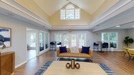 Located at 100 Eclipse Dr in Cary, NC, this single-story community clubhouse offers a thoughtfully designed gathering space with a variety of functional areas. The centerpiece of the building is a grand great room distinguished by soaring vaulted ceilings with an arched transom window that floods the space with natural light. French doors line the exterior walls, opening views to the surrounding greenery and providing seamless indoor-outdoor connectivity. Rich hardwood flooring runs throughout the main living areas, anchored by stylish area rugs and contemporary furnishings including tufted leather and upholstered sofas, accent chairs, and curated décor. Crown molding and a two-tone wall treatment lend a polished, classic character to the space. Adjacent to the great room is a dedicated conference and media room equipped with a wall-mounted flat-screen TV, a long communal table, and vibrant yellow upholstered seating — ideal for meetings or presentations. Large windows in this room maintain the bright, open atmosphere. A kitchenette area features white cabinetry with granite countertops and large windows overlooking the landscaped exterior. Additional nook seating areas with garden views provide quiet retreats throughout the facility, making this clubhouse a versatile and welcoming community hub.