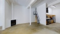 This single-floor space showcases a raw, industrial aesthetic defined by polished concrete floors and bright white-painted walls and ceiling. Exposed structural beams run across the ceiling, adding architectural character while reinforcing the loft-style character of the space. A central white support column anchors the open-plan layout, which offers a generous, flexible footprint suitable for a variety of uses.

A standout feature is the sleek black metal ship ladder, which leads to a mezzanine or loft platform above, providing additional vertical space without occupying significant floor area. This industrial-style ladder serves as both a functional element and a design focal point within the room.

On one wall, a fireplace adds a contrasting touch of warmth and homeliness to the otherwise minimalist industrial environment. To the right, cabinetry is partially visible, suggesting a kitchen or utility area. Natural light filters into the space, enhancing the clean, open atmosphere created by the all-white surfaces. Overall, the space combines utilitarian design with practical features, making it a highly adaptable environment.