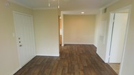 This single-floor space offers a clean, functional layout designed with everyday comfort in mind. The main living area is finished with rich, dark hardwood-style flooring that runs throughout, adding warmth and visual depth to the interior. Neutral, light beige walls provide a versatile backdrop that complements a variety of furnishing styles and décor choices.

The living area benefits from a combination of recessed ceiling lighting and pendant fixtures, offering layered illumination that enhances the open feel of the room. Crown molding along the ceiling adds a subtle architectural detail that elevates the overall finish of the space.

A partial wall and open passageway lead into the kitchen area, which is visible in the background and features wooden cabinetry, maintaining a cohesive warm tone throughout the home. A wall-mounted ventilation register is also present, indicating a central HVAC system for year-round climate control.

The layout flows naturally, with clean transitions between spaces supported by white-trimmed doorways and baseboards. The overall ambiance is bright, tidy, and move-in ready, making it a practical and welcoming space suitable for a range of occupants.