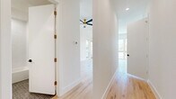 This thoughtfully designed single-floor unit offers a clean, modern aesthetic throughout, highlighted by light-toned hardwood floors and crisp white walls that create a bright, airy atmosphere. The open living area is a standout feature, anchored by a sleek ceiling fan with gold hardware and flooded with natural light from a generous bank of windows that frame outdoor views. The vaulted ceiling adds an elevated sense of volume to the space.

The kitchen is efficiently laid out with white cabinetry, white countertops, and a full suite of stainless steel appliances including a refrigerator, electric range, and over-the-range microwave. A tall window brings in additional natural light, enhancing the clean, functional design. The adjacent hallway connects the main living areas seamlessly, with recessed lighting and a smart thermostat adding to the unit's modern conveniences.

The bathroom is elegantly appointed with a floating white vanity accented by a gold bar pull, an undermount sink, a large mirror, and gold-toned faucet fixtures. The tub and shower combination features textured white tile surround, while the floor is finished with charcoal hexagonal mosaic tile, providing a sophisticated contrast. A matte black door handle adds a refined finishing touch to this well-curated space.