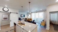 This single-floor unit showcases a thoughtfully designed open-concept layout where the kitchen, dining, and living areas flow seamlessly together. The kitchen is a standout feature, equipped with stainless steel appliances, a full suite of white cabinetry, subway tile backsplash, and a generous granite-topped island with a double sink and pendant lighting above. Bar seating along the island adds casual dining flexibility, while the adjacent dining area provides a dedicated space for meals.

The living room benefits from large sliding glass doors dressed in sheer curtains, filling the space with natural light and offering outdoor access. Light wood-tone flooring runs throughout, complementing the neutral wall palette. A sliding barn door adds a contemporary farmhouse touch, connecting the main living area to adjoining spaces.

The bedroom is a calm, well-proportioned retreat featuring a ceiling fan, natural light from a window with blinds, and a closet. The room connects directly to a bathroom and opens toward the main living area, reinforcing the unit's cohesive, open feel. Ceiling fans are present in both the living room and bedroom, contributing to overall comfort throughout this well-finished, modern space.