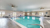 This generously sized indoor pool facility offers a bright, well-appointed aquatic environment designed for both recreation and function. The centerpiece of the space is a large, rectangular swimming pool with clear turquoise water, framed by a wide, wood-look tile deck that provides ample room for poolside seating and circulation on all sides. The facility is contained within a single, open floor plan that maximizes usable space and promotes an airy atmosphere.

Natural light flows generously throughout the room via numerous large, double-hung windows positioned along multiple walls, complemented by recessed ceiling lighting and multiple ceiling fans that ensure comfortable air circulation year-round. The walls are finished in neutral tones with blue accent walls near the entrance area, lending the space a clean and calming aesthetic.

A set of glass-paned French doors serves as the main entry point, opening to views of the surrounding community. An accessible pool lift is present, along with handrail-equipped pool steps, emphasizing the facility's commitment to inclusive use. Safety equipment, including life rings and emergency signage, is strategically placed throughout. A windowed pass-through or viewing area connects to an adjacent room, adding to the facility's functional design. The overall ambiance is clean, well-lit, and welcoming.
