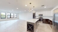 This contemporary single-floor residence showcases a thoughtfully designed open-concept layout that seamlessly integrates the living and kitchen areas. The herringbone-patterned light wood flooring flows throughout the main living space, creating a warm yet sophisticated aesthetic. Recessed lighting and large black-framed windows allow natural light to fill the generously proportioned living area, which also features an exposed ductwork detail that adds an industrial-modern touch.

The kitchen is a standout centerpiece, outfitted with two-tone cabinetry — high-gloss white upper cabinets paired with deep espresso lower cabinets — complemented by white quartz countertops and a matching backsplash. A large island with built-in wine storage anchors the space, while a pair of sculptural pendant lights add elegance overhead. Stainless steel appliances complete the culinary suite.

The bedroom opens directly to a private balcony through sliding glass doors, offering outdoor views of the surrounding neighborhood and abundant natural light, with a ceiling fan adding comfort.

The bathrooms are finished to a high standard, featuring large-format marble-look porcelain tile, a floating dual-sink vanity with backlit mirrors, and a glass-enclosed shower, combining clean lines with a luxurious ambiance throughout.