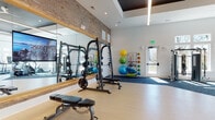 This single-level fitness center offers a thoughtfully designed workout environment that balances industrial character with modern functionality. The space is anchored by a striking exposed brick accent wall, which contrasts with crisp white walls and a coffered ceiling detail featuring linear lighting. The elevated ceiling height contributes to an open, airy atmosphere throughout.

The facility is divided into distinct workout zones, each with purpose-appropriate flooring. Light hardwood-style flooring covers the free weights and strength training area, where a Smith machine, weight bench, and plate-loaded equipment are positioned alongside large wood-framed mirrors with integrated TV displays. Blue carpet tiles define the functional training and cardio zones, housing TRX suspension systems, medicine ball racks, resistance machines, and stability balls on organized wall-mounted racks.

A dedicated cardio section features Matrix treadmills positioned directly in front of an expansive bank of tall windows, flooding the space with natural light and providing unobstructed outdoor views. Additional cardio equipment, including a stationary bike, is positioned nearby. Double glass doors and a side entry door further enhance natural light and accessibility. The overall design is clean, modern, and highly functional.