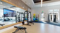 This single-level fitness center offers a thoughtfully designed workout environment that balances industrial character with modern functionality. The space is anchored by a striking exposed brick accent wall, which contrasts with crisp white walls and a coffered ceiling detail featuring linear lighting. The elevated ceiling height contributes to an open, airy atmosphere throughout.

The facility is divided into distinct workout zones, each with purpose-appropriate flooring. Light hardwood-style flooring covers the free weights and strength training area, where a Smith machine, weight bench, and plate-loaded equipment are positioned alongside large wood-framed mirrors with integrated TV displays. Blue carpet tiles define the functional training and cardio zones, housing TRX suspension systems, medicine ball racks, resistance machines, and stability balls on organized wall-mounted racks.

A dedicated cardio section features Matrix treadmills positioned directly in front of an expansive bank of tall windows, flooding the space with natural light and providing unobstructed outdoor views. Additional cardio equipment, including a stationary bike, is positioned nearby. Double glass doors and a side entry door further enhance natural light and accessibility. The overall design is clean, modern, and highly functional.