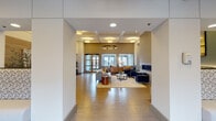 This single-floor amenity space is thoughtfully designed to offer a well-rounded communal experience, combining comfort, functionality, and contemporary style. The centerpiece of the space is a spacious lounge area distinguished by soaring ceilings with coffered detailing, a striking circular pendant light fixture, and a sleek floor-to-ceiling tile fireplace. Plush seating arrangements in navy blue and gray, anchored by a soft area rug, create an inviting atmosphere bathed in natural light from large windows. Adjacent to the lounge is a cozy nook with teal accent walls, a live-edge shelf, and additional seating beside the fireplace surround. The kitchen and café-style dining area features a sweeping curved stone countertop, warm pendant lighting, wood-paneled walls, and banquette seating, making it ideal for casual gatherings. Two dedicated conference rooms provide professional meeting spaces, each furnished with a large white table and ergonomic mesh chairs, with one room offering natural light through slatted blinds. The well-equipped fitness center rounds out the amenities, housing a comprehensive array of cardio machines, cable systems, and strength equipment, all set against mirrored walls and accented with teal wall tones throughout.