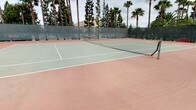 This outdoor recreational facility offers two well-maintained sports courts designed for active use and community engagement. The basketball court features a regulation backboard and hoop, with clearly marked court lines on a terracotta-toned hard surface. Padded pole guards provide added safety, and the court is fully enclosed by a tall chain-link fence with mesh windscreen panels, ensuring both security and reduced wind interference during play. A gated entry allows easy access to the court area.

Adjacent to the basketball court is a full-sized tennis court, surfaced in a classic combination of green and terracotta hard court material with crisp white boundary lines and a centered net in place. The court's enclosure mirrors the same chain-link and mesh fencing design, maintaining a cohesive and polished appearance throughout the facility.

Both courts are set against a lush backdrop of tall, mature palm trees that lend the space a tropical, resort-inspired character. The surrounding greenery provides a degree of natural shade and visual privacy. The overall environment is open-air, benefiting from natural light and outdoor ambiance, making this facility an inviting destination for tennis players, basketball enthusiasts, and recreational users alike.