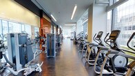 This fitness centre offers a thoughtfully designed single-floor layout that combines functionality with a clean, modern aesthetic. The space features an elongated open floor plan, allowing for an organized arrangement of equipment without feeling crowded. A row of cardio machines — including ellipticals, stair climbers, and stationary bikes — lines one side of the facility, while strength and resistance training equipment occupies the opposing side, creating clear pathways for easy navigation throughout the space.

The interior is finished with dark hardwood-style flooring that contrasts sharply with bright white walls and ceilings, contributing to a polished, professional atmosphere. Linear LED lighting runs along the ceiling, providing consistent, even illumination throughout. Large floor-to-ceiling windows at the far end of the space flood the facility with natural light and offer views of the surrounding urban environment, enhancing the overall workout experience.

Mirrored wall panels are incorporated along sections of the space, visually expanding the area while serving a practical purpose for users monitoring their form. The overall ambiance is clean, well-maintained, and contemporary, making it a professional-grade environment suited for a comprehensive fitness routine.