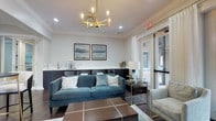 This single-floor space presents a thoughtfully designed interior that balances comfort and contemporary style. The lounge area serves as the centerpiece, anchored by a plush teal velvet sofa accented with patterned throw pillows and set atop a warm, grid-patterned area rug. A striking gold geometric chandelier with exposed candelabra bulbs hangs overhead, casting a refined glow that complements the recessed lighting and crisp crown molding throughout. The walls are painted in a soft, neutral tone and adorned with a pair of framed landscape artwork, contributing to a calm and curated ambiance. A built-in refreshment station lines the back wall, featuring dark cabinetry, a countertop appliance, and a small sink — ideal for hosting or everyday convenience. Natural light filters generously through large sliding glass doors dressed with sheer white curtains, opening to an outdoor area and enhancing the airy feel of the room. Additional seating, including a light upholstered accent chair, offers flexible arrangements for conversation or relaxation. The combination of rich wood-toned flooring, gold metallic accents, and layered lighting creates a polished, inviting atmosphere suited for both casual gatherings and quiet moments.