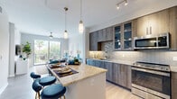 This well-designed one-bedroom, one-bathroom residence in Doral, Florida offers a thoughtfully organized open-concept layout across a single floor. The kitchen is a standout feature, showcasing wood-grain cabinetry, stainless steel appliances including a refrigerator, range, microwave, and dishwasher, a tile backsplash, and a granite-topped island with seating — ideal for casual dining. Track lighting and elegant pendant lights illuminate the space, which flows seamlessly into the living area. The living room is bright and airy, benefiting from large sliding glass doors that open to a private balcony with outdoor views of lush tropical greenery. A ceiling fan adds comfort to the space. The bedroom is generously proportioned, featuring large windows framing views of palm trees, plush carpet flooring, and a ceiling fan. The bathroom is well-appointed with a granite vanity, wood-tone cabinetry, a large walk-in glass shower with tile surround, and light tile flooring. Light wood-style flooring runs through the main living areas, adding warmth throughout. A dedicated closet and hallway complete the functional floor plan of this modern Doral residence.