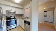 This single-floor residence offers a practical and cohesive layout connecting several well-defined living areas. The kitchen is efficiently designed with white shaker-style cabinetry, granite-look countertops, and a mosaic tile backsplash that adds visual texture. Stainless steel appliances, including an electric range and dishwasher, complement the space, while light wood-look flooring ties the kitchen seamlessly to the adjacent living area. A structural half-wall partially separates the kitchen, maintaining an open feel while defining each zone. The living area is a standout space, featuring the same wood-look flooring, neutral gray walls, and a full-width sliding glass door that opens to a private balcony with outdoor views and ample natural light. The bedroom offers plush carpeting, neutral tones, and a window that brings in additional daylight. The bathroom includes a single-sink vanity with a granite-look countertop, a tub-shower combination with white tile surround, and matching wood-look flooring. A washer and dryer unit is also visible within the residence, adding everyday convenience to the overall functional design of the space.