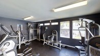 This single-level fitness room is designed to support a well-rounded workout routine. The space is finished with dark charcoal walls that create a focused, gym-like atmosphere, complemented by rubber flooring that provides durability and safety underfoot. Overhead fluorescent lighting ensures the room is well-lit throughout.

The room is outfitted with a multi-function cable strength training machine, a seated resistance machine, a rowing or leg press apparatus, a set of dumbbells, and what appears to be a cardio machine along the far wall. This selection of equipment caters to both strength training and cardiovascular fitness.

A row of windows along one wall allows natural light to filter into the space, with views of an outdoor patio area featuring seating and surrounding greenery. Additional clerestory windows near the ceiling further enhance the brightness of the room while maintaining privacy. The combination of practical equipment, durable finishes, and natural light makes this a functional and inviting space for regular fitness use.