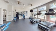 Located at 3415 SW 39th Blvd in Gainesville, Florida, this well-appointed fitness center offers a comprehensive workout facility across a single floor. The space is divided into distinct functional zones, each designed to support a full range of exercise routines.

The main workout area is bright and open, featuring durable rubber flooring, recessed lighting, and ceiling fans that maintain comfortable airflow. Large windows along one wall flood the space with natural light while offering views of an outdoor pool area, creating an energizing atmosphere. Cardio equipment, including multiple treadmills, a stationary bike, and an elliptical, is positioned to take advantage of these views. A wall-mounted television adds entertainment options for those on cardio machines.

A dedicated strength training section houses a cable machine system and a multi-station weight machine with adjustable benches, all accompanied by bold abstract artwork that adds a touch of style to the functional space.

A separate room serves as a free weights and functional training area, equipped with a comprehensive rack of kettlebells, medicine balls, and a wall-length mirror to assist with form and technique. An adjustable bench and diamond-plate flooring detail complete this zone.

Additional amenities include restroom facilities with ADA-accessible features, ensuring the space is welcoming to all users. Crown molding and clean white walls throughout lend the facility a polished, well-maintained appearance.