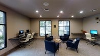This single-floor amenity space is thoughtfully designed to serve as both a functional business center and a welcoming lounge area. The room features neutral-toned walls and a smooth ceiling equipped with recessed lighting, which ensures consistent, well-distributed illumination throughout the space. Patterned wall-to-wall carpeting in soft, neutral tones adds texture underfoot and contributes to the room's refined yet approachable aesthetic.

A dedicated workstation area lines one wall, complete with a floating desk surface, desktop computers, and ergonomic white office chairs — ideal for residents or guests who need a quiet place to work or study. Adjacent to this, a built-in floating shelf extends along the wall, providing additional workspace or surface area.

Large black-framed windows dominate the exterior wall, flooding the interior with natural light and offering pleasant views of the surrounding greenery outside. The lounge area is centered around a set of deep navy blue accent chairs arranged around a round wooden coffee table, creating an inviting setting for casual conversation or relaxed reading. A ceiling-mounted speaker and HVAC vent reflect the space's attention to comfort and functionality. Overall, the space strikes a balance between professional utility and comfortable design.