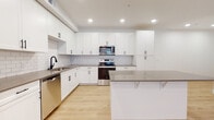 This single-floor unit showcases a cohesive modern design highlighted by light hardwood-style flooring, crisp white walls, and black hardware accents throughout. The kitchen serves as a central feature, outfitted with full-height white shaker cabinetry, a white subway tile backsplash, and grey quartz countertops. A peninsula provides additional workspace and casual seating. Stainless steel appliances include a range with microwave, dishwasher, and refrigerator, complemented by a deep undermount sink with a matte black faucet. Recessed lighting illuminates the space effectively.

The living area offers a clean, open layout with light wood flooring, recessed lighting, and a glass-panel exterior door flanked by windows fitted with horizontal blinds, allowing natural light to enter. Bedrooms feature neutral carpet, windows with blinds, and generous walk-in closets equipped with double hanging rods and shelving.

Bathrooms are finished with white shaker vanities, quartz countertops, and black hardware. The primary bath includes a dual-sink vanity, a glass-enclosed shower, and direct access to a walk-in closet. The secondary bath features a full tub-shower combination. The overall aesthetic is clean, contemporary, and move-in ready.
