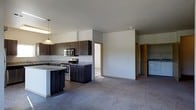 This single-floor space showcases a thoughtfully designed open-concept layout that seamlessly connects the kitchen and living area. The kitchen is a standout feature, equipped with dark wood cabinetry that extends to the upper walls, providing generous storage. Granite countertops add a polished, durable work surface, while stainless steel appliances — including a range and microwave — offer a sleek, modern aesthetic. A kitchen island with seating provides additional prep space and a casual dining option, illuminated by pendant lighting overhead.

The living area flows naturally from the kitchen, finished with plush neutral carpeting that adds warmth and comfort underfoot. Crisp white walls and a clean, minimalist design allow natural light to brighten the space. A doorway visible from the main living area leads to additional rooms beyond, suggesting a practical and well-organized floor plan. In the background, a laundry area with a washer is visible, adding everyday convenience. The overall ambiance is contemporary and functional, with a cohesive neutral palette of dark cabinetry, light counters, and soft grey carpeting creating a balanced and inviting interior.