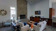 This single-story home showcases a striking living room as its centerpiece, defined by a floor-to-ceiling stacked stone fireplace that commands immediate attention. The fireplace surround, finished in layered natural stone, extends the full height of the soaring vaulted ceiling, creating a dramatic architectural focal point. Large windows along the adjacent wall allow ample natural light to fill the space, enhancing the airy, open atmosphere.

The living area is thoughtfully arranged with comfortable neutral-toned sofas and armchairs accented by rich blue throw pillows, centered around a rectangular wood coffee table. A warm wood media console with built-in shelving and cabinetry sits opposite the fireplace, providing both display and storage space alongside a wall-mounted flat-screen television. Decorative accessories, including ceramic vases and accent lamps, add character throughout the room.

The cool gray walls complement the warm stone tones of the fireplace, while the overall layout balances both style and functionality. The high ceilings and open design contribute to a spacious, welcoming ambiance suited for both relaxed daily living and entertaining.