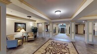 This building's interior showcases a thoughtfully designed common area that combines classic architectural elements with a welcoming, functional layout. The main lobby serves as the focal point of the space, featuring decorative columns with ornate capitals framing open passageways, crown molding along the ceilings, and a combination of tile and patterned area rugs underfoot. Recessed lighting and flush-mount fixtures illuminate the space evenly, while wall sconces add a warm, ambient glow.

A grand staircase with rich mahogany-toned wood banisters and balusters, fitted with patterned carpet treads, connects the levels and serves as a striking architectural centerpiece. An open upper landing with matching railings overlooks the lower common areas, and large potted plants placed throughout add a refreshing natural element.

The expansive lower-level hallways are finished with soft patterned carpeting and feature multiple doorways leading to additional rooms. Wainscoting and paneled wall details run throughout, lending a refined, traditional character. A formal entry with dark wood double doors and sidelights provides a distinguished point of arrival. Overall, the space conveys a welcoming, institutional elegance suited for a communal residential or hospitality setting.