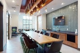 This thoughtfully designed collaborative space offers multiple distinct zones suited for both group gatherings and focused work. The centerpiece of the main area is a substantial live-edge wood conference table surrounded by dark upholstered chairs, anchored on a deep green area rug. Overhead, an eye-catching suspended wood beam structure adorned with Edison bulb pendant lights adds warmth and industrial character to the soaring ceiling height. A full accent wall clad in textured teal ceramic tile serves as a striking backdrop for a wall-mounted flat-screen television, flanked by warm wood cabinetry with a built-in sink and dark countertop — ideal for catering or refreshments. Large steel-framed windows flood the space with natural light and offer views of the surrounding streetscape. A separate private meeting room, enveloped in deep teal walls, features a matte dark conference table with upholstered seating and a floor-to-ceiling window that draws in abundant daylight. Additional casual seating nooks with counter-height tables and pendant lighting provide flexible options for smaller gatherings or independent work throughout the space.