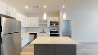 Located at 3 Gilbert in Merrimack, New Hampshire, this single-floor residence offers a thoughtfully designed layout with modern finishes throughout. The kitchen serves as a central highlight, featuring crisp white cabinetry with sleek bar-pull hardware, light quartz countertops, and a full suite of stainless steel appliances including a refrigerator, range, over-the-range microwave, and dishwasher. A contrasting dark-toned island with pendant lighting above adds both visual interest and additional workspace, while light wood-look flooring ties the kitchen and adjacent living area together seamlessly. The open living and dining area benefits from the same durable hardwood-style flooring and receives natural light through generously sized windows. Bedrooms are finished with plush wall-to-wall carpeting and feature windows that bring in natural light, creating comfortable, well-lit retreats. The bathroom includes a full soaking tub and shower combination with a clean white surround, complemented by a wood-toned vanity with modern hardware and a well-lit mirror. A hallway connects the private spaces efficiently, with white panel doors and soft neutral wall tones maintaining a cohesive, contemporary aesthetic throughout the entire residence.
