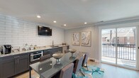 This single-floor clubhouse offers a well-appointed, modern gathering space designed for both community events and everyday resident use. The main area seamlessly blends a dining space and kitchenette, anchored by a sleek glass-top dining table surrounded by upholstered chairs atop a vivid teal-accented area rug. The kitchenette features dark cabinetry with granite countertops, a white subway tile backsplash, a stainless steel sink, a built-in microwave, and a wall-mounted flat-screen television — making it fully functional for social gatherings. Recessed lighting and light hardwood-style flooring run throughout, contributing to a bright and welcoming atmosphere. Large sliding glass doors and a glass panel door open to an outdoor balcony, allowing natural light to fill the space and providing views of the surrounding community. A leasing office area is distinguished by a bold teal accent wall and features a central work desk, comfortable lounge chairs, and curated wall art, lending a professional yet inviting feel. A wide, well-lit corridor connects the various areas of the clubhouse, lined with artwork and leading to additional access points. The overall design palette of soft grays, teal, and warm wood tones creates a cohesive, contemporary ambiance throughout.