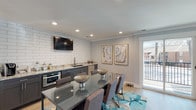 This single-floor clubhouse offers a well-appointed, modern gathering space designed for both community events and everyday resident use. The main area seamlessly blends a dining space and kitchenette, anchored by a sleek glass-top dining table surrounded by upholstered chairs atop a vivid teal-accented area rug. The kitchenette features dark cabinetry with granite countertops, a white subway tile backsplash, a stainless steel sink, a built-in microwave, and a wall-mounted flat-screen television — making it fully functional for social gatherings. Recessed lighting and light hardwood-style flooring run throughout, contributing to a bright and welcoming atmosphere. Large sliding glass doors and a glass panel door open to an outdoor balcony, allowing natural light to fill the space and providing views of the surrounding community. A leasing office area is distinguished by a bold teal accent wall and features a central work desk, comfortable lounge chairs, and curated wall art, lending a professional yet inviting feel. A wide, well-lit corridor connects the various areas of the clubhouse, lined with artwork and leading to additional access points. The overall design palette of soft grays, teal, and warm wood tones creates a cohesive, contemporary ambiance throughout.