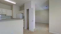 This single-floor residence presents an open and functional layout that seamlessly connects the kitchen and living areas. The kitchen is outfitted with white cabinetry, a white refrigerator, and a countertop peninsula that serves as a natural divider between the cooking and living spaces. Light tile flooring grounds the kitchen area, providing a practical and easy-to-maintain surface.

A six-panel white door opens to reveal a built-in closet featuring shelving and hanging rods, offering convenient storage solutions. This organizational feature is centrally positioned, making it accessible from the main living space.

The living area extends generously beyond the kitchen, finished with neutral-toned carpet that adds warmth underfoot. The walls are painted in soft, light tones that contribute to an airy and bright atmosphere throughout. Visible HVAC vents in the ceiling suggest a consistent climate control system integrated into the space.

Overall, the residence maintains a clean, understated aesthetic with a cohesive neutral color palette, practical storage, and a straightforward open-plan design that maximizes usability across the shared living and kitchen zones.