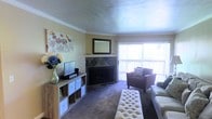 This single-floor space centers around a well-appointed living room that balances comfort and functionality. The room's most prominent feature is a tile-surround fireplace with a diamond-pattern design, topped by a framed wall mirror that adds depth and visual interest. Crown molding lines the ceiling, lending a polished, finished look to the warm yellow walls. Soft, plush carpeting runs throughout, contributing to the room's comfortable atmosphere. A large sliding glass door fitted with vertical blinds allows generous natural light to pour in, brightening the entire space. The layout accommodates a variety of furnishing arrangements, as evidenced by a tufted ottoman serving as a central coffee piece, a cushioned armchair positioned near the window, and a low-profile media console with cube storage along one wall. Decorative touches, including floral artwork and fresh blooms in a glass vase, add a welcoming, lived-in quality. Overall, the space offers a cozy yet functional environment suited to everyday living and relaxed entertaining.