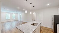 Located at 11122 Hambright Junction Drive in Huntersville, NC, this well-appointed single-floor residence offers three bedrooms, two bathrooms, and a thoughtfully designed open layout. The kitchen is a standout feature, showcasing crisp white cabinetry reaching to the ceiling, granite countertops, stainless steel appliances including a range and built-in microwave, and a large island with a sink — all set atop warm hardwood-style flooring that flows throughout the home. Recessed lighting and pendant fixtures keep the space bright and functional. The living area connects seamlessly to the kitchen, creating an inviting open-concept environment. The bedrooms are bright and airy, with several featuring transom-topped doors opening to a private balcony that overlooks a lush, tree-lined backdrop — providing a serene natural setting. The primary bedroom includes direct access to an en-suite bathroom. Both bathrooms are finished with granite-topped vanities and clean white tile, with one featuring a full tub-shower combo accented by subway tile. Closets are equipped with wire shelving systems for organized storage. The home's neutral palette, consistent flooring, and modern hardware give it a cohesive, move-in-ready aesthetic throughout.