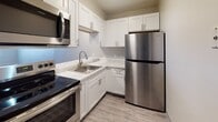 This single-floor residence offers a thoughtfully designed layout that balances functionality and comfort. The kitchen is outfitted with crisp white shaker-style cabinetry, light quartz countertops, and a full suite of stainless steel appliances, including a refrigerator, electric range, over-the-range microwave, and dishwasher. A neutral color palette and wood-look vinyl flooring give the kitchen a clean, contemporary feel.

An open pass-through counter connects the kitchen to the main living area, creating an airy, connected flow between spaces. The living area is finished with the same wood-look vinyl flooring and features flush-mount ceiling lighting. A hallway leads to the additional rooms of the residence.

A spacious living or sitting room is carpeted in a warm neutral tone and features a large sliding glass door with vertical blinds, allowing natural light to fill the space and providing access to an outdoor area. A wall-mounted air conditioning unit serves this room.

A separate carpeted bedroom offers a quiet retreat with ample wall space. The bathroom features a full soaking tub with a shower and is accented by large-format marble-look ceramic wall tiles with elegant veining, complemented by chrome fixtures, lending a polished and refined aesthetic to the space.