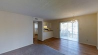 This single-floor space offers an open and functional layout that connects the living, dining, and kitchen areas seamlessly. The living area features plush carpeting, creating a comfortable and inviting atmosphere, while the adjoining dining and kitchen zones transition to hardwood-style flooring for a clean, practical finish. A textured ceiling adds visual character throughout.

The kitchen is positioned behind a half-wall pass-through, providing a sense of openness while maintaining a defined cooking space. It features light-toned cabinetry, a built-in range, and a countertop that doubles as a casual serving area. A hallway beyond the kitchen suggests additional rooms, offering privacy from the main living space.

Natural light flows generously into the space through large sliding glass doors, which open onto a private balcony enclosed with decorative lattice railings and views of surrounding greenery. An elegant chandelier above the dining area adds a refined touch and enhances the overall brightness. The neutral color palette of white walls and warm flooring tones creates a cohesive, welcoming ambiance throughout.