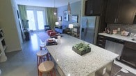 This single-floor residence showcases an open-concept design that seamlessly integrates the kitchen and living areas into one cohesive, flowing space. The kitchen is a standout feature, anchored by a generous granite-topped island with a speckled finish that provides both prep space and casual seating for multiple bar stools. Dark espresso cabinetry lines the kitchen walls, complemented by stainless steel appliances, including a French door refrigerator, creating a sleek, contemporary aesthetic.

Pendant lighting with elongated metallic fixtures hangs above the island, adding a modern decorative touch while providing focused illumination. The open layout transitions naturally into the living area, which is furnished with a blue sofa, colorful accent seating, and layered area rugs atop polished concrete flooring. The blue-toned walls lend a calm, cohesive ambiance throughout.

Natural light pours in generously through oversized French doors dressed with floor-to-ceiling green drapes, which open to an exterior space and contribute to the airy, open feel of the residence. The overall atmosphere is modern, vibrant, and thoughtfully designed for comfortable everyday living.