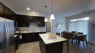 This single-floor space showcases a thoughtfully designed open-concept layout that seamlessly connects the kitchen and dining areas. The kitchen is appointed with rich, dark espresso cabinetry that extends to the upper and lower walls, providing ample storage while creating a striking visual contrast against the light-colored countertops and neutral gray walls. A central kitchen island with a bright white surface serves as both a functional workspace and a casual gathering spot, illuminated by two modern pendant lights that hang at varying heights, adding an elegant focal point to the space. Recessed lighting throughout ensures the kitchen remains well-lit. The cabinetry includes a built-in dishwasher and a sink positioned along the perimeter counter. The open layout flows naturally into the dining area, which accommodates a full dining table and chairs, bathed in natural light from a large window fitted with vertical blinds. Decorative wall art enhances the dining space, adding personality to the neutral surroundings. Hardwood-style flooring runs throughout, tying the two areas together cohesively and contributing to a clean, contemporary ambiance.
