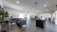 This single-story space presents an open-plan layout that seamlessly integrates multiple functional zones within one expansive area. The interior is finished with wide-plank wood-look flooring in a cool grey tone, complementing the neutral white walls and creating a clean, cohesive aesthetic throughout.

A reception or concierge desk finished in dark granite anchors one side of the space, equipped with a computer workstation and decorative greenery. Adjacent to it, a comfortable lounge area features a contemporary grey sofa paired with a sleek glass coffee table, ideal for relaxed seating or informal gatherings.

Moving deeper into the space, café-style high-top tables with bar stools in a soft blue-grey tone provide additional seating options. Natural light filters in through windows dressed with sheer curtains, brightening the far end of the room. Colorful framed artwork adorns the walls, adding visual interest without overwhelming the understated palette.

Recessed lighting and a ceiling fan are integrated overhead, ensuring the space remains well-lit and comfortable. Overall, the design strikes a balance between professional functionality and welcoming comfort.