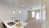 Located at 2323 Wolf Ranch Pkwy in Georgetown, Texas, this single-floor residence offers a thoughtfully designed open-concept layout that promotes a sense of spaciousness and ease of movement throughout. The kitchen serves as a central focal point, showcasing a clean white countertop with a sleek undermount sink and a modern arc faucet. Elegant cylindrical pendant lights hang above the counter, adding a contemporary touch to the space. The kitchen flows naturally into the living area, which is finished with plush carpeting that adds warmth and comfort underfoot. A ceiling fan with integrated lighting anchors the living space, contributing to both function and comfort. Natural light filters in through multiple windows dressed with classic white blinds, brightening the interior and creating an inviting atmosphere. A doorway at the far end of the living area provides direct access to the outdoors, enhancing the sense of openness. The neutral color palette of soft whites and warm beiges throughout creates a calm, versatile backdrop well-suited for a variety of interior styles. Overall, this residence combines practical design with modern detailing for comfortable everyday living.
