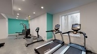 Located at 422 E Boston Post Rd in Mamaroneck, NY, this fitness center offers a well-appointed, single-floor exercise facility designed to support a comprehensive workout experience. The space is finished with a clean, modern aesthetic, featuring crisp white walls accented by bold teal feature walls that energize the environment. Durable grey flooring runs throughout, providing a practical and polished surface suited for a range of fitness activities.

The cardio zone is equipped with multiple TRUE-brand treadmills and a rowing-style machine, each featuring integrated digital displays for performance tracking. Natural light filters in through a window along one wall, adding a bright and welcoming atmosphere to the cardio area. The strength and functional training section houses a multi-station cable machine, an adjustable weight bench, a full rack of hex dumbbells, TRX suspension straps, and exercise mats — offering versatile options for both resistance and bodyweight training.

Recessed ceiling lighting provides even illumination throughout the facility, complemented by a wall-mounted split HVAC unit ensuring year-round climate comfort. A dedicated restroom area features a sleek, modern floating vanity with a chrome-framed base, a rectangular vessel sink with a contemporary faucet, and a well-lit mirror — offering a clean and functional space for post-workout freshening up.