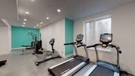 Located at 422 E Boston Post Rd in Mamaroneck, NY, this fitness center offers a well-appointed, single-floor exercise facility designed to support a comprehensive workout experience. The space is finished with a clean, modern aesthetic, featuring crisp white walls accented by bold teal feature walls that energize the environment. Durable grey flooring runs throughout, providing a practical and polished surface suited for a range of fitness activities.

The cardio zone is equipped with multiple TRUE-brand treadmills and a rowing-style machine, each featuring integrated digital displays for performance tracking. Natural light filters in through a window along one wall, adding a bright and welcoming atmosphere to the cardio area. The strength and functional training section houses a multi-station cable machine, an adjustable weight bench, a full rack of hex dumbbells, TRX suspension straps, and exercise mats — offering versatile options for both resistance and bodyweight training.

Recessed ceiling lighting provides even illumination throughout the facility, complemented by a wall-mounted split HVAC unit ensuring year-round climate comfort. A dedicated restroom area features a sleek, modern floating vanity with a chrome-framed base, a rectangular vessel sink with a contemporary faucet, and a well-lit mirror — offering a clean and functional space for post-workout freshening up.