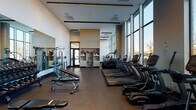 This contemporary fitness center is designed to support a full range of workout routines in a bright, professionally appointed environment. The space is organized into distinct functional zones, each thoughtfully laid out to maximize usability and flow. Floor-to-ceiling windows line one side of the facility, flooding the interior with natural light and offering open views of the surrounding area, creating an energizing atmosphere for exercise. The cardio zone features a row of TRUE-brand treadmills positioned directly in front of the expansive windows, alongside elliptical trainers, a rowing machine, and a stationary spin bike. The strength training area is anchored by a full rack of dumbbells in graduated weights, accompanied by adjustable benches and a cable machine, with large wall-mounted mirrors reflecting the space and allowing users to monitor their form. Dark tile flooring extends throughout the main areas, while rubber flooring underlies the free weight section for added safety and durability. The ceilings are notably high, contributing to a spacious, open feel. A built-in wooden shelving unit near the entrance adds a warm, refined touch, and frosted-glass sliding doors define a secondary training area, offering a degree of separation for focused workouts. The overall design balances modern aesthetics with practical functionality.